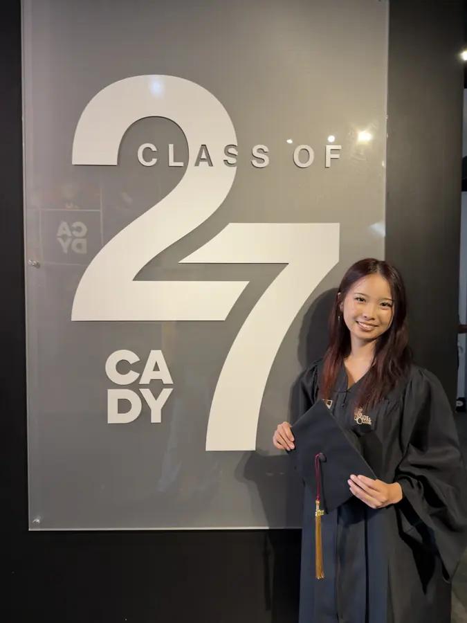 Korrine Lee in a graduation cap and gown beside a large Class of 27 sign